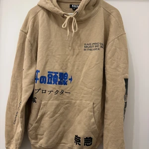 Beige hoodie från William Baxter M - Säljer en beige hoodie från William Baxter med coola japanska och engelska tryck i blått och svart. Tröjan har huva med snörning, känguruficka och långa ärmar. Perfekt för dig som gillar streetwear och unika detaljer.