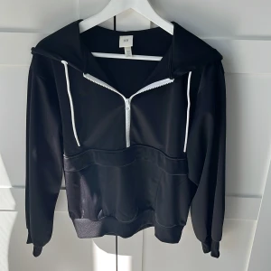 Svart hoodie med half zip från H&M - Svart hoodie från H&M med vit half zip-dragkedja och vita snören i huvan. Tröjan har en stor ficka framtill med dragkedja och är tillverkad i ett slätt, syntetiskt material. Långärmad och med ribbade muddar för en clean look.