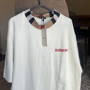 Vit Burberry t-shirt med logga XL - Vit t-shirt från Burberry i oversized passform med klassisk rund hals i beige, svart, vit och röd. Broderad röd Burberry-logga på bröstet. Tillverkad i mjuk bomull och har korta ärmar. Perfekt för dig som gillar stilrena och lyxiga plagg.