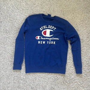 Blå Champion sweatshirt med tryck - Blå sweatshirt från Champion med stort vitt och rött tryck på bröstet där det står ATHL.DEPT. och New York. Tröjan har rund hals, långa ärmar och ribbade muddar. Perfekt för en sportig och avslappnad stil.