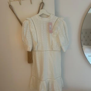 Vit broderad kortklänning med volang - Superfin vit kortklänning i bomull med broderade blommor, volangdetaljer och puffärmar. Klänningen har knappar vid ärmslut, markerad midja och v-ringning i ryggen. Perfekt för sommarens alla tillfällen och lappen kvar! 