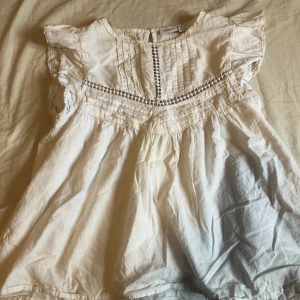 Vit blus med spets från Name It - Supersöt vit blus från Name It med volangärmar och fina spetsdetaljer längs axlar och bröst. Blusen har rund halsringning, dekorativa sömmar och ett luftigt fall. Perfekt för dig som gillar romantisk och boho-inspirerad stil.