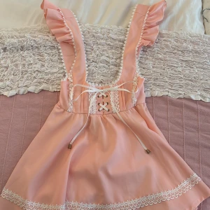 Söt rosa kortklänning med spets - Supergullig ljusrosa kortklänning med volangprydda axelband, snörning framtill och vita spetsdetaljer längs kanterna. Kjolen är utsvängd och har extra volang nertill. Perfekt för dig som gillar en romantisk och lekfull stil. Den är i storlek S men har stretch så den sitter inte super tight!! <3