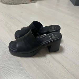 Svarta klackade sandaler från Vox - Svarta sandaler från Vox med bred rem över foten, öppen tå och chunky klack. Skorna har plattformsula och är tillverkade i syntetmaterial. Perfekta för dig som gillar en stilren och edgy look.