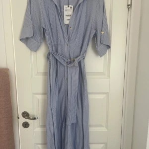 Randig midi-klänning från Zara - Snygg blåvit midi-klänning från Zara med tunna vertikala ränder, knappar framtill och matchande bälte i midjan. Klänningen har krage, korta ärmar med knappdetalj och är perfekt för en fräsch sommarlook.