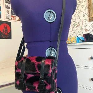 Dr. Martens axelväska i rosa och svart fuskpäls - Cool axelväska från Dr. Martens med fluffig rosa och svart leopardmönstrad/komönstrad fuskpäls på framsidan och slät svart baksida i skinn. Väskan har justerbar axelrem och snygga spänndetaljer framtill. Perfekt för klubben eller en dag på stan. I princip helt ny, köpt för en månad sen och använd ett fåtal gånger. 