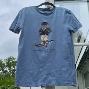 Blå Polo Bear t-shirt från Ralph Lauren - Snygg blå t-shirt från Polo Ralph Lauren med ikoniskt Polo Bear-tryck där björnen åker skidor. Modellen är custom slim fit och har rund hals och korta ärmar. Perfekt för dig som gillar klassisk stil med en twist. Använd typ 5 gånger