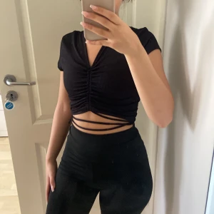 Svart croppad topp från Divided XS - Svart croppad topp från Divided i ribbad bomull med kort ärm, v-ringning och snygg snörning som kan knytas runt midjan. Toppen har rynkade detaljer framtill som ger en cool look. Perfekt till höga byxor eller kjol.