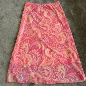 Rosa långkjol med paisleymönster - Snygg långkjol i rosa med paisleyinspirerat mönster i ljusa toner av gult, lila och vitt. Kjolen har en rak passform och resår i midjan för extra komfort. Perfekt för sommaren