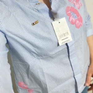 Randig skjorta med broderade blommor - Snygg blå- och vitrandig skjorta från U.S. Polo Assn. med broderade rosa blommor på bröstet och ärmen. Skjortan har klassisk krage, knappar framtill och trekvartsärm. Perfekt för dig som vill ha en skjorta med lite extra detaljer.