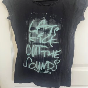 Svart t-shirt med tryck - Svart t-shirt med coolt graffiti-inspirerat tryck i ljusblått på framsidan där det står 'LET'S KICK OUT THE SOUNDS'. T-shirten har korta ärmar och en klassisk rund halsringning. Materialet känns mjukt och passar perfekt till en avslappnad streetstil. Finns ingen lapp men uppskattar strlk S