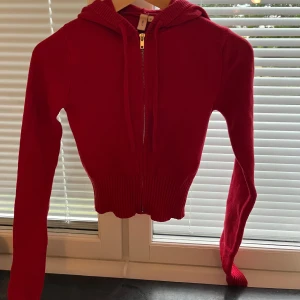 Röd stickad hoodie från Nelly - Snygg röd stickad hoodie från Nelly i storlek S. Tröjan är croppad med ribbad nederkant, har huva och dragkedja framtill. Långärmad modell.