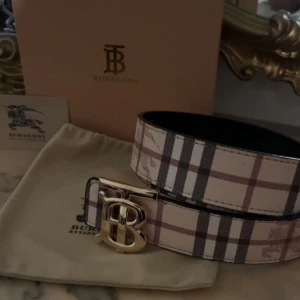 Burberry rutigt bälte med guldspänne - Reverse snyggt bälte från Burberry med klassiskt rutigt mönster i beige, brunt och svart. Bältet har ett stort guldigt 'TB'-spänne framtill och är gjort i skinn. Perfekt accessoar för att lyfta din outfit med en lyxig touch. Du kan byta från beige till svarta sidan.
