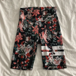 Blommiga träningstights-shorts från Stronger - Snygga högmidjade cykelshorts från Stronger i svart med blommigt mönster i rosa, rött och grönt. Perfekta för träning och har två coola vita ränder på ena benet.