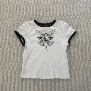 Vit t-shirt med svart tryck  - Vit t-shirt från Divided med svarta kontrastkanter vid hals och ärmslut. Framsidan har ett stort svart tryck av en fe med texten 'Always Love'. Modellen är kortärmad och har en klassisk passform. Nästan aldrig använd