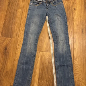 Blå lågmidjade jeans med guld-detaljer - Säljer ett par blå jeans med snyggt slitna detaljer och coola guld-broderier samt paljetter på bakfickorna. Jeansen har låg midja, klassisk femficksmodell och raka ben. Perfekta för dig som vill sticka ut lite extra med detaljer på fickorna.