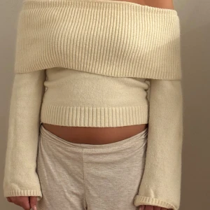 Offshoulder stickad tröja i beige - Superfin offshoulder stickad tröja i ljusbeige med bred ribbad kant upptill och vid midjan. Tröjan har långärmad passform och sitter tajt över kroppen, perfekt för dig som gillar en trendig och stilren look.