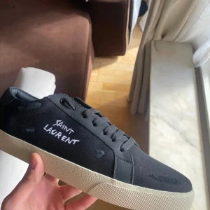 Svarta Saint Laurent sneakers - Snygga svarta sneakers från Saint Laurent med vit broderad logga på sidan. Skorna har en klassisk låg profil, svarta snören och detaljer i både textil och skinn. Yttersulan är beige och ger en schysst kontrast till den svarta ovandelen. Allt Og medföljer, billig för en anledning.