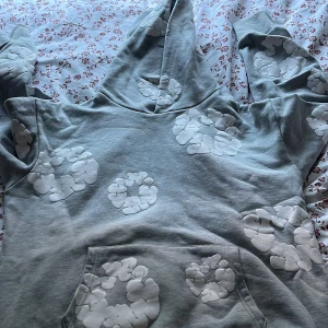 Grå hoodie från Denim Tears med blommor - Säljer en grå hoodie från Denim Tears med vita blommiga tryck över hela tröjan. Tröjan har huva, magficka och långa ärmar. Tillverkad i mjuk bomull, perfekt för dig som gillar streetwear med unik design. Använd endast 2-3 gånger, så anses som 10/10 skick!