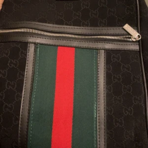Gucci ryggsäck med GG-mönster - Säljer en svart Gucci ryggsäck med klassiskt GG-monogram över hela väskan. Framsidan har gröna och röda ränder och silverfärgade dragkedjor med Gucci-logga. Väskan är i textil med detaljer i skinn och har flera fack och justerbara axelremmar.