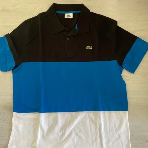 Lacoste pikétröja i svart, blå och vit - Snygg pikétröja från Lacoste Sport med färgblock i svart, blått och vitt. Klassisk krage, korta ärmar och broderad krokodil-logga på bröstet. Perfekt för dig som gillar sportig och stilren vibe. Tillverkad i mjuk bomull för skön känsla hela dagen.