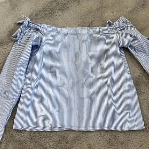 Randig offshoulder blus från H&M - Säljer en blåvit randig offshoulder blus från H&M i 100% bomull. Blusen har långa ärmar med knytband vid axlarna och en luftig, rak passform. Perfekt för dig som gillar en fräsch och somrig stil med snygga detaljer.