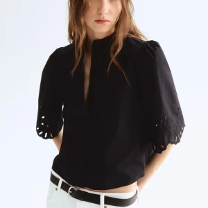 Svart blus med broderade ärmar - Svart blus i bomull med vida, halvlånga ärmar och broderade detaljer samt hålmönster vid ärmsluten. Rak passform, liten ståkrage och knäppning framtill. Perfekt för dig som gillar unika detaljer och stilren look.