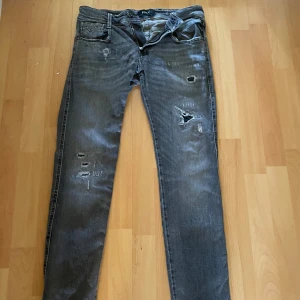 Replay Anbass grå slitna jeans - Säljer ett par grå jeans från Replay, modell Anbass. Jeansen har slim fit passform och är snyggt slitna med flera hål och slitningar på benen. Klassisk femficksmodell med dragkedjegylf. Perfekta för en cool och avslappnad stil.