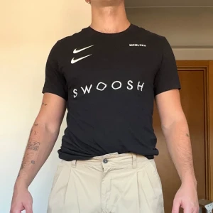 Nike t-shirt med Swoosh-tryck - Svart t-shirt från Nike med vit Swoosh-logga både fram och bak samt texten 'SWOOSH' på bröstet. Klassisk passform och rund halsringning. Tillverkad i mjuk bomull, perfekt för en sportig och avslappnad stil.