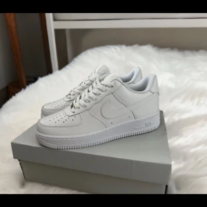 Nike Air Force 1 helvita sneakers - Klassiska Nike Air Force 1 sneakers i helvitt skinn. Snygg och clean design med perforerad tå, rund tå och platt sula. Lågt skaft och vita snören. Perfekta för dig som gillar stilrena och tidlösa sneakers med streetkänsla.