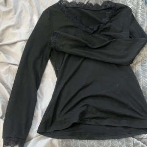 Svart långärmad topp med spetsdetaljer - Svart långärmad topp med v-ringning och fina spetsdetaljer vid halsringning och ärmslut. Snygg och enkel modell som sitter tajt mot kroppen. Perfekt att styla med jeans eller kjol för en clean look. Aldrig använd 