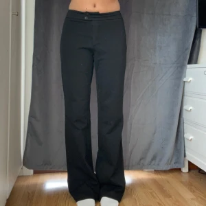 Svarta vintage kostymbyxor från Zoul, strl 36 - Svarta kostymbyxor från Zoul i storlek 36. Byxorna har hög midja, raka ben med lätt bootcut och stängs med två knappar framtill. Materialet är en mix av polyester och viskos med lite elastan för extra komfort. Har ett litet hål precis under gylfen, går enkelt att laga med lite tråd!!