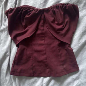 Vinröd bandeau tubtopp med volang - Supersnygg vinröd bandeau tubtopp med volangdetaljer och lätt rynk framtill. Toppen är axelbandslös och har en figurnära passform som framhäver siluetten. Perfekt för dig som vill sticka ut med en cool och trendig look.