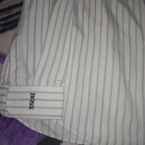 Vit randig skjorta från BOSS - Säljer en klassisk vit skjorta från BOSS med tunna blå ränder och broderad logga på bröstet. Skjortan har knappar framtill och krage, perfekt för en clean och stilren look.