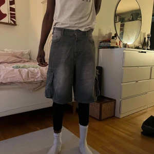 Grå cargoshorts med fickor - Säljer ett par grå cargoshorts med stora sidofickor och lös passform. Shortsen har klassisk jeanslook och är knälånga, perfekta för en avslappnad stil.