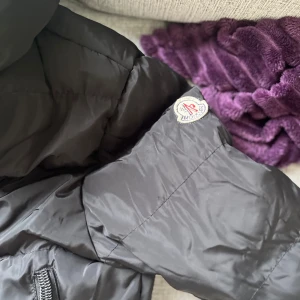 Svart dunjacka från Moncler - Säljer en svart dunjacka från Moncler med huva och två dragkedjeförsedda fickor framtill. Jackan har klassisk pufferdesign och Moncler-logga på ärmen. Perfekt för kyliga dagar och riktigt snygg till streetwear-stilen.