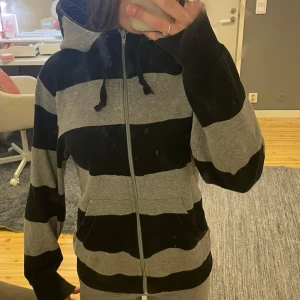 Svart och grå randig hoodie med dragkedja - Säljer en svart och grå hoodie med breda ränder och dragkedja framtill. Tröjan har huva med snören och två fickor fram. Har inte hunnit tvätta den ännu så det kan finnas någon fläck men jag tvättar den såklart innan jag fraktar 🤗Jag vet inte vad det är för märke.