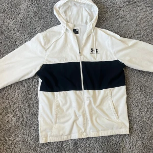 Vit och svart vindjacka från Under Armour - Snygg vit vindjacka med svart panel över bröstet från Under Armour. Jackan har huva, dragkedja framtill och två sidofickor. Perfekt för blåsiga dagar och sportiga outfits.