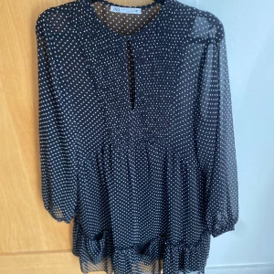 Svart prickig klänning från Zara, stl S - Svart klänning från Zara med vita prickar och långärmade transparenta ärmar. Klänningen har volangdetalj nertill och en lätt v-ringning framtill. Perfekt för dig som gillar en lekfull och trendig stil.