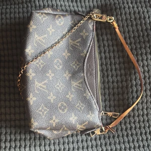 Monogram axelväska - Säljer en klassisk stilfull axelväska i brunt monogrammönster med guldfärgade detaljer och kedja. Väskan har dragkedja och röd insida. Perfekt accessoar för dig som gillar lyxiga detaljer och tidlös design.