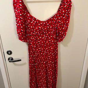 Röd mönstrad klänning med puffärmar - Röd klänning med vita och svarta detaljer, puffiga kortärmar och rundad halsringning. Klänning stängs med en dragkedja i sidan och har även slits vid sidan. Väldigt fin på! Använd 1 gång.
