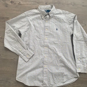 Ralph Lauren skjorta  - Klassisk vit skjorta med små blå och beige rutor från Ralph Lauren. Skjortan har lång ärm, button-down krage och broderad logga på bröstet. Perfekt för en stilren och tidlös look.