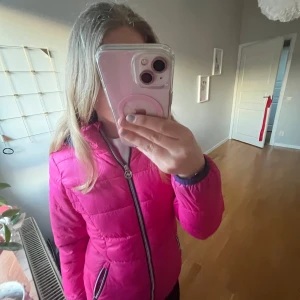 Rosa pufferjacka med huva från Michael kors - Säljer en snygg rosa pufferjacka från Woxer med svart dragkedja och huva. (Michael kors)Jackan har långa ärmar och två fickor framtill med dragkedja. Perfekt för kyligare dagar och ger en poppig färg till outfiten.