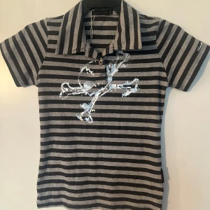 Randig piké t-shirt med tryck fr criminal damage S/M - Snygg kortärmad piké t-shirt från Calvin Klein i svart och grått med breda ränder och ett stort vitt tryck framtill. Klassisk krage och knäppning vid halsen. Perfekt för en avslappnad stil. Helt ny. Från Criminal damage. S/M.