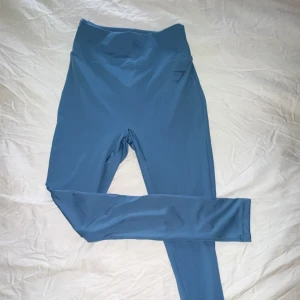 Blå träningstights från Gymshark - Säljer ett par blå träningstights från Gymshark med hög midja och diskret logga på höften. Tightsen har en tight passform och är tillverkade i ett stretchigt syntetmaterial. Baksidan har scrunch-detalj för extra form.