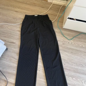 Randiga kostymbyxor  - Säljer ett par svarta kostymbyxor från NA-KD med smala vita ränder. Byxorna har hög midja och raka ben med klassisk passform. Perfekta för en stilren look och passar till många olika outfits.