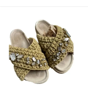  Inuikii tofflor  - Unika sandaler med breda, stickade remmar i olivgrönt och dekorerade med stora glittrande stenar. Skorna har en platt, ljus sula och öppen tå. Perfekta för dig som vill sticka ut med en trendig och bekväm look.