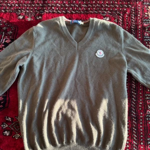 Olivgrön stickad tröja från Moncler, Vintage - Säljer en olivgrön stickad tröja från Moncler med v-ringad hals och broderad Moncler-logga på bröstet. Tröjan har långa ärmar och klassisk passform, Vintage moncler fint skick!