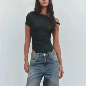 grön body zara - Säljer en mörkgrön bodysuit med oneshoulder-design och draperad effekt. Toppen har en tight passform och är tillverkad i ett stretchigt material som sitter snyggt på kroppen. Perfekt att styla med jeans eller kjol.
