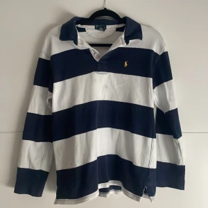 Randig pikétröja från Polo Ralph Lauren - Klassisk långärmad pikétröja från Polo Ralph Lauren med breda marinblå och vita ränder. Tröjan har krage, knäppning vid halsen och den ikoniska broderade loggan i gult på bröstet. Perfekt för en sportig och avslappnad stil.